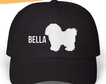 May include: Black baseball cap featuring a white silhouette of a dog and the name "BELLA" in white block letters. The text "EMBROIDERED CAN BE PERSONALIZED" is above the cap's brim. A selection of color options are displayed.