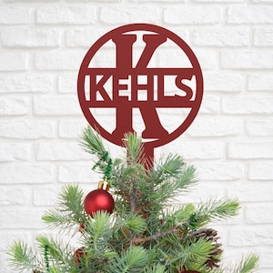May include: A burgundy metal Christmas tree topper with the name "KEHLS" in a circular design. The topper is placed on a small evergreen tree decorated with a red ornament and green garland.