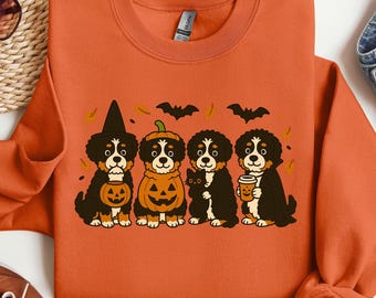 May include: Orange sweatshirt featuring a Halloween-themed graphic. The design includes four Bernese Mountain Dogs dressed in costumes, including a witch hat, pumpkin, and holding a coffee cup. Bats and leaves are also included.