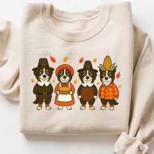 May include: Beige sweatshirt featuring a Thanksgiving-themed design. The graphic shows four Bernese Mountain Dogs dressed as pilgrims and a farmer, surrounded by autumn leaves. The dogs are wearing hats, aprons, and holding a pie and corn.