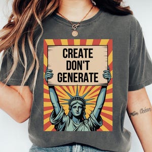 May include: A charcoal gray t-shirt featuring a graphic print of the Statue of Liberty holding a sign that reads CREATE DON'T GENERATE. The background of the graphic consists of red and yellow sunburst rays, and the shirt has a crew neckline.