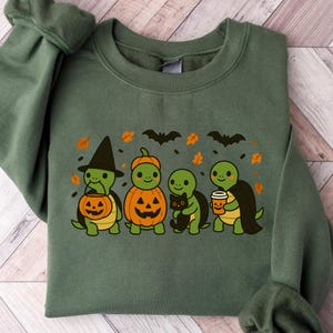 May include: Green sweatshirt featuring a Halloween-themed design. The graphic shows four cartoon turtles dressed in costumes: a witch, a pumpkin, a turtle holding a black cat, and a turtle with a coffee cup. Bats and leaves complete the design.
