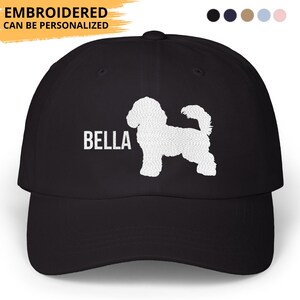 May include: Black baseball cap featuring a white silhouette of a dog and the name "BELLA" in white block letters. The text "EMBROIDERED CAN BE PERSONALIZED" is above the brim. The cap has a curved brim and a fabric strap closure.