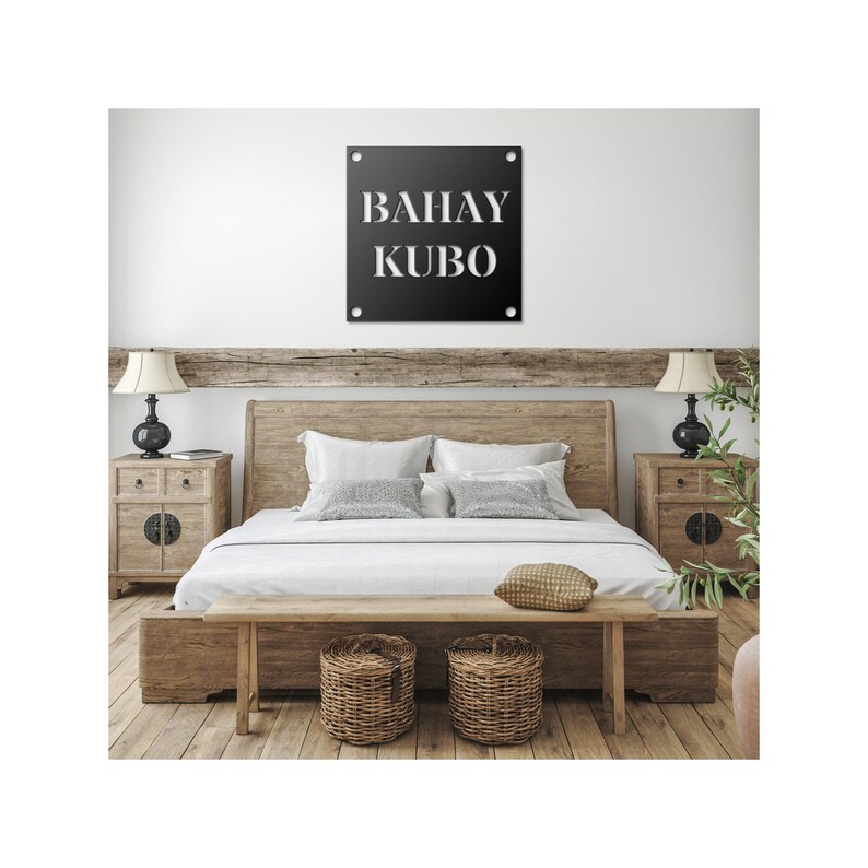 May include: A black metal sign with white lettering that reads "BAHAY KUBO" hanging on a white wall above a wooden bed with white linens and a wooden bench.