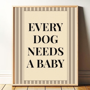 May include: A framed print with the text "EVERY DOG NEEDS A BABY" in black, centered on a cream-colored background. The frame is light brown, and the print has a striped border in shades of beige and brown.
