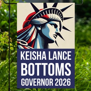 May include: A garden flag featuring a stylized image of the Statue of Liberty in red, white, and blue. The flag has the text "KEISHA LANCE BOTTOMS GOVERNOR 2026" in white letters on a navy blue background. The flag is displayed outdoors.