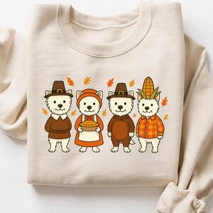 May include: Beige sweatshirt featuring a Thanksgiving-themed graphic. The design shows four cartoon dogs dressed in festive attire, including pilgrim hats, aprons, and a corn husk. Autumn leaves are scattered around the dogs.