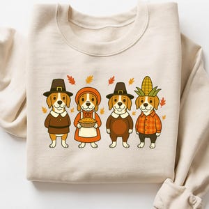 May include: Beige sweatshirt featuring a Thanksgiving-themed design. Four cartoon beagle dogs are dressed in pilgrim and harvest attire, including hats, dresses, and a corn husk. Autumn leaves are scattered around the dogs.