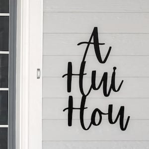 May include: Black metal sign with the words "A Hui Hou" in a cursive font. The sign is mounted on a white, horizontal-planked wall next to a white door frame and a window.