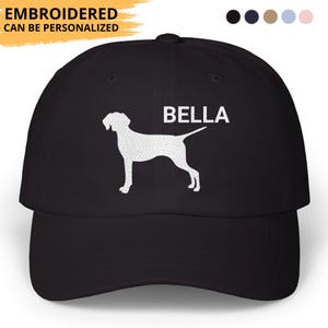 May include: Black baseball cap with a white silhouette of a dog and the name "BELLA" printed on the front. The text "EMBROIDERED CAN BE PERSONALIZED" is above the brim. The cap has a curved brim and a button on top.