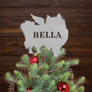 May include: A silver metal dog silhouette with the name "BELLA" cut out, placed on a small Christmas tree with red ornaments and pinecones. The background is a dark brown wood.