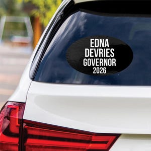 May include: An oval-shaped black car decal applied to a rear vehicle window. The sticker features bold white sans-serif text that reads EDNA DEVRIES GOVERNOR 2026, arranged in four stacked lines centered within the black oval background.