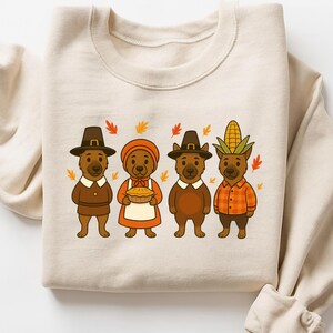 May include: Beige sweatshirt featuring a Thanksgiving-themed graphic. The design includes four cartoon bears dressed in pilgrim and harvest attire, surrounded by autumn leaves. The bears wear hats and hold a pie and corn.