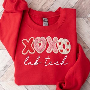 May include: A bright red sweatshirt with the embroidered words "XOXO lab tech". The letters X and O are red, the heart is pink, and the O has small red hearts. The sweatshirt is folded.