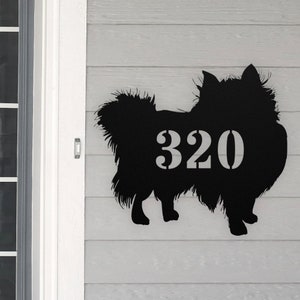 May include: Black metal silhouette of a Pomeranian dog with the house number 320 cut out of the dog's body. The silhouette is mounted on a light gray wooden surface.
