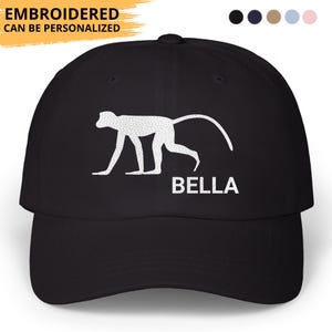 May include: Black baseball cap featuring a white monkey silhouette and the name "BELLA" in white. The text "EMBROIDERED CAN BE PERSONALIZED" is above the cap's brim. The cap has a curved brim and a classic six-panel design.