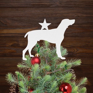 May include: White silhouette of a dog with a star on top, used as a Christmas tree topper. The dog is on top of a green Christmas tree with red ornaments.