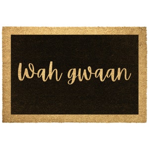 May include: Rectangular doormat with a black center and a gold border. The words "Wah gwaan" are written in a gold, cursive font across the center of the mat. The doormat is made of a natural fiber material.