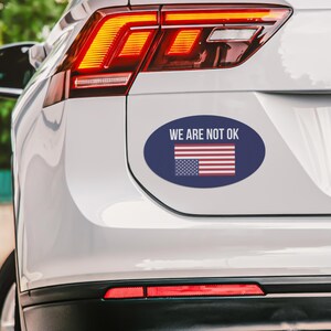 May include: A white car with a blue oval sticker that says "WE ARE NOT OK" with an upside-down American flag.