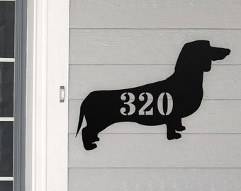 May include: Black metal silhouette of a dachshund with the house number 320 cut out of the dog's body. The silhouette is mounted on a light gray wooden surface.