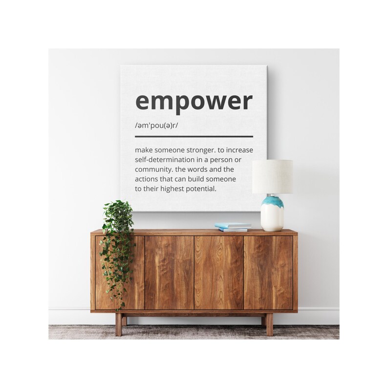 May include: A white canvas print with the word "empower" in black text and a definition of the word below. The print is hanging on a white wall above a wooden cabinet with a potted plant and a lamp.
