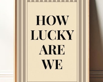 May include: Framed wall art with the text "HOW LUCKY ARE WE" in bold black letters. The artwork has a cream-colored background with a black border and is set within a wooden frame. The frame has a striped pattern in shades of beige and brown.