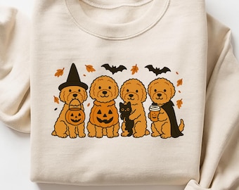 May include: Cream-colored sweatshirt featuring a Halloween-themed graphic. The design includes four cartoon dogs dressed in costumes: a witch, a pumpkin, a black cat, and a coffee cup. Bats and leaves complete the festive scene.