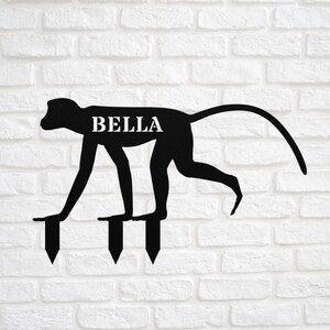 May include: Black metal monkey silhouette garden stake with the name "BELLA" on the body. The stake has three metal spikes for inserting into the ground.