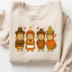 May include: Beige sweatshirt featuring a Thanksgiving-themed design. The graphic shows four cartoon bears dressed in festive attire, including pilgrim hats and a corn husk headdress. Autumn leaves are scattered around the bears. The sweatshirt is made of a soft material.