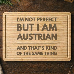 May include: A rectangular wooden cutting board with a carved border. The text "I'M NOT PERFECT BUT I AM AUSTRIAN AND THAT'S KIND OF THE SAME THING" is engraved in the center. The board has a light brown color and a natural wood grain.