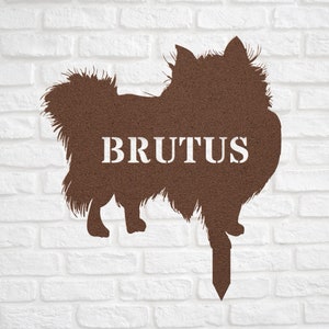 May include: Brown metal silhouette of a dog with the name "BRUTUS" written in white on the body. The silhouette is attached to a metal stake for outdoor use.