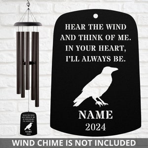 May include: Black metal wind chime hanger with a white raven silhouette and the text "Hear the wind and think of me. In your heart, I'll always be."  The hanger also includes a space for a name and year.  "NAME 2024" is shown as an example.  "Wind chime is not included" is printed below the hanger.
