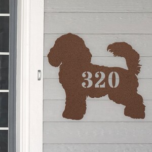 May include: A brown metal silhouette of a dog with the house number 320 cut out of the silhouette. The silhouette is mounted on a light gray wooden wall.