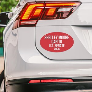 May include: A red oval bumper sticker with white text that reads "SHELLEY MOORE CAPITO U.S. SENATE 2026." The sticker is affixed to the rear of a white vehicle, near the taillight.