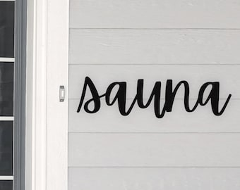 Könnte beinhalten: Schwarzes Metallschild mit dem Wort "Sauna" in kursivschrift. Das Schild ist an einer weißen Holzwand befestigt.