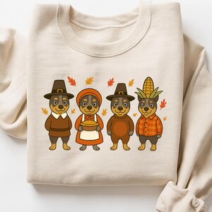 May include: Beige sweatshirt featuring a Thanksgiving-themed design. The graphic shows four cartoon dogs dressed in festive attire, including pilgrim hats and a corn husk headdress. Autumn leaves are scattered around the dogs.