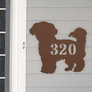 May include: A brown metal silhouette of a dog with the house number 320 cut out of the silhouette. The dog is mounted on a light gray wooden wall.