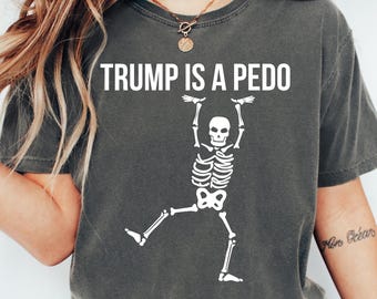 May include: Dark gray t-shirt with the white text "TRUMP IS A PEDO" above a white skeleton graphic. The skeleton is holding onto the text with its arms and has one leg raised. The shirt has short sleeves.