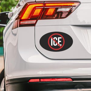 May include: A black oval sticker with the text "ICE" in white, crossed out by a red circle and line. The sticker is affixed to the rear of a white vehicle, near the taillight.
