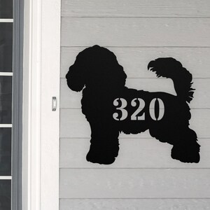 May include: Black metal silhouette of a dog with the house number 320 cut out of the silhouette. The silhouette is mounted on a light gray wooden surface.