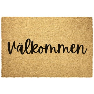 May include: A rectangular doormat made of natural coir fiber. The mat features the word "Välkommen" in a black, cursive font, centered on the mat. The background is a light brown color.