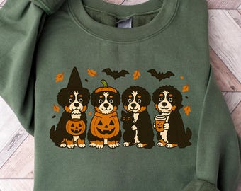 May include: Green sweatshirt featuring a Halloween-themed design. The graphic shows four Bernese Mountain Dogs dressed in costumes, including a witch hat, pumpkin, and holding a coffee cup. The design includes bats and fall leaves.