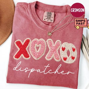 May include: A dusty rose t-shirt with the word "XOXO" in a knitted design, featuring a red X, a pink heart, a pink X, and a cream-colored O with red hearts. The word "dispatcher" is written in cursive below the design.