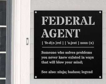 May include: Black metal sign with the words "FEDERAL AGENT" in bold white letters. The sign includes a definition and the phrases "ninja; badass; legend". It is mounted on a white wall.