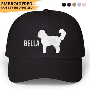 May include: Black baseball cap featuring a white silhouette of a dog and the name "BELLA" in white block letters. The text "EMBROIDERED CAN BE PERSONALIZED" is above the cap's brim. The cap has a curved brim and a fabric strap closure.