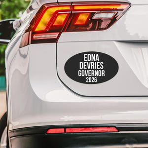 May include: An oval-shaped black car sticker with white text that reads EDNA DEVRIES GOVERNOR 2026. The sticker is applied to the rear trunk of a white vehicle, positioned below the red taillight assembly and above the rear bumper reflector.