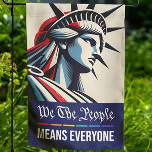 May include: A garden flag featuring a stylized illustration of the Statue of Liberty in red, white, and blue. The flag has the text "We The People MEANS EVERYONE" in white on a navy blue banner with a rainbow stripe.