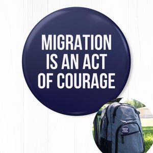 May include: A navy blue button with white text that reads "MIGRATION IS AN ACT OF COURAGE." A backpack with the same button attached is in the lower right corner. The button is round and has a glossy finish.