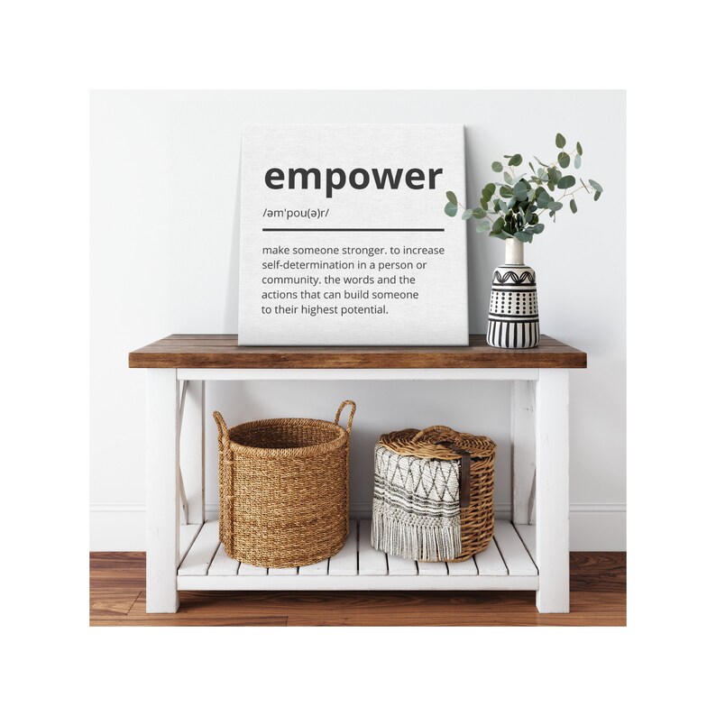 May include: A white canvas print with the word "empower" in black text and a definition of the word. The print is sitting on a white console table with a brown top. There are two woven baskets on the bottom shelf of the table and a vase with a green plant on the top shelf.