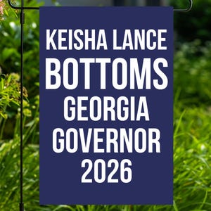 May include: A navy blue garden flag with white text. The flag reads "KEISHA LANCE BOTTOMS GEORGIA GOVERNOR 2026". The flag is displayed outdoors in front of a green background.